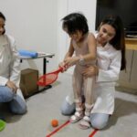 Duas mulheres de jaleco branco ajudam uma criança a se movimentar em um escritório com uma raquete de tênis de plástico nas mãos.