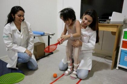 Duas mulheres de jaleco branco ajudam uma criança a se movimentar em um escritório com uma raquete de tênis de plástico nas mãos.