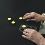 Mãos de uma pessoa idosa manuseiam peças táteis circulares amarelas sobre uma mesa preta. À frente, há uma base com recortes circulares, onde as peças podem ser encaixadas.