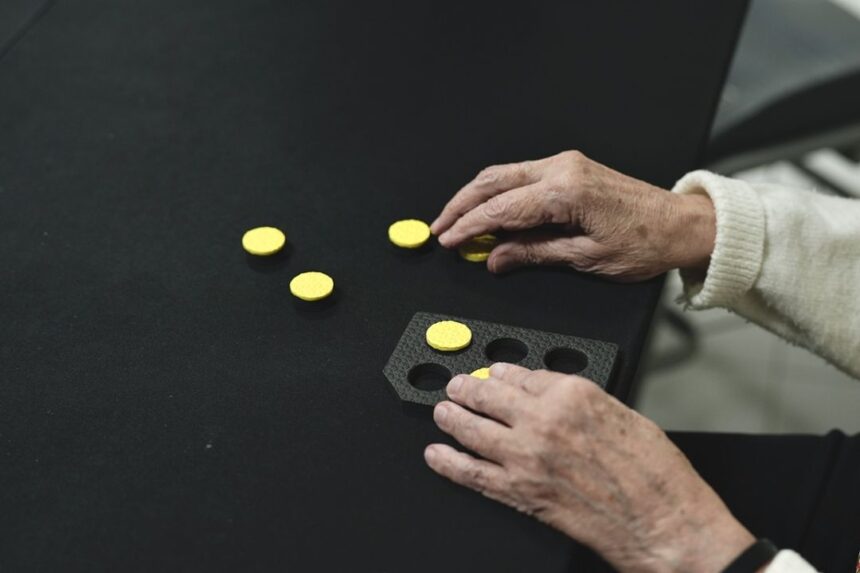 Mãos de uma pessoa idosa manuseiam peças táteis circulares amarelas sobre uma mesa preta. À frente, há uma base com recortes circulares, onde as peças podem ser encaixadas.