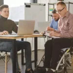 Três homens estão em uma sala de escritório moderna, trabalhando juntos. Dois estão sentados em cadeiras comuns e um deles, que usa cadeira de rodas.