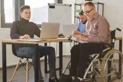 Três homens estão em uma sala de escritório moderna, trabalhando juntos. Dois estão sentados em cadeiras comuns e um deles, que usa cadeira de rodas.