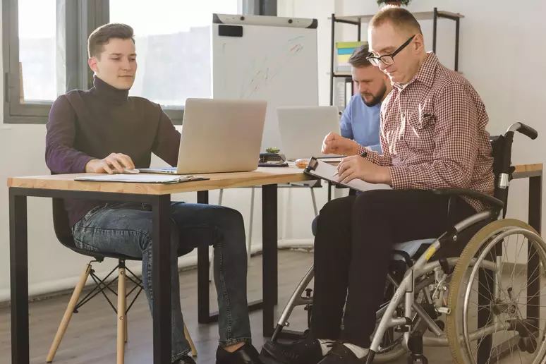 Três homens estão em uma sala de escritório moderna, trabalhando juntos. Dois estão sentados em cadeiras comuns e um deles, que usa cadeira de rodas.