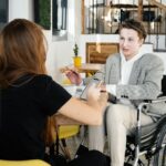 Duas pessoas conversam em um ambiente interno moderno, semelhante a uma cafeteria. Uma delas, uma mulher jovem, está de costas, segurando um celular. A outra pessoa, que está de frente, é um homem jovem cadeirante, vestindo blazer xadrez, moletom branco e calça clara. Ele segura uma caneta e parece estar explicando algo, enquanto há um caderno aberto sobre a mesa. Ao fundo, vasos pequenos com plantas decoram o local.