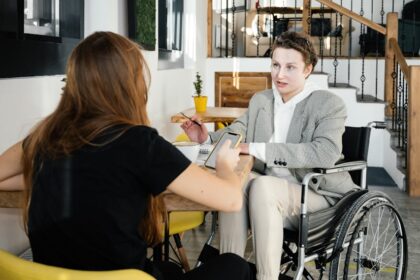 Duas pessoas conversam em um ambiente interno moderno, semelhante a uma cafeteria. Uma delas, uma mulher jovem, está de costas, segurando um celular. A outra pessoa, que está de frente, é um homem jovem cadeirante, vestindo blazer xadrez, moletom branco e calça clara. Ele segura uma caneta e parece estar explicando algo, enquanto há um caderno aberto sobre a mesa. Ao fundo, vasos pequenos com plantas decoram o local.