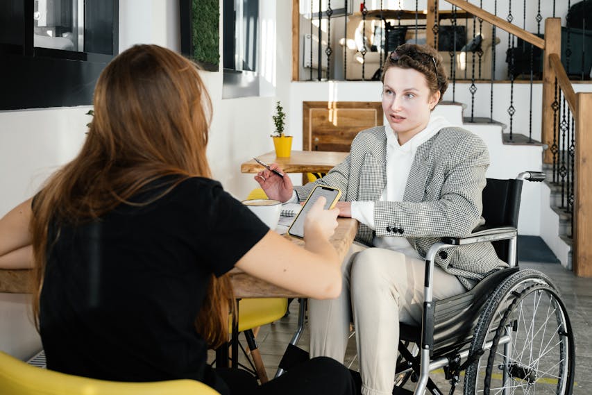 Duas pessoas conversam em um ambiente interno moderno, semelhante a uma cafeteria. Uma delas, uma mulher jovem, está de costas, segurando um celular. A outra pessoa, que está de frente, é um homem jovem cadeirante, vestindo blazer xadrez, moletom branco e calça clara. Ele segura uma caneta e parece estar explicando algo, enquanto há um caderno aberto sobre a mesa. Ao fundo, vasos pequenos com plantas decoram o local.
