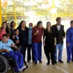 Grupo diverso de pessoas, incluindo pessoas com deficiência, posa sorridente para uma foto em um espaço esportivo com estrutura amarela ao fundo.