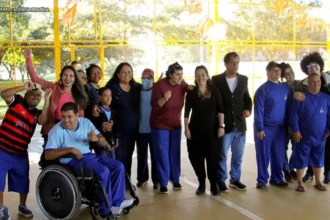 Grupo diverso de pessoas, incluindo pessoas com deficiência, posa sorridente para uma foto em um espaço esportivo com estrutura amarela ao fundo.