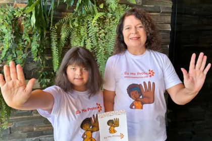 Uma mulher adulta e uma menina com síndrome de Down estão em pé, lado a lado, sorrindo e erguendo a mão espalmada à frente, em sinal de 'pare'. Ambas vestem camisetas brancas com a frase 'Eu Me Protejo' e a ilustração de uma menina com a mão levantada em sinal de proteção.