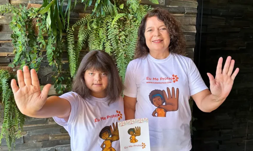 Uma mulher adulta e uma menina com síndrome de Down estão em pé, lado a lado, sorrindo e erguendo a mão espalmada à frente, em sinal de 'pare'. Ambas vestem camisetas brancas com a frase 'Eu Me Protejo' e a ilustração de uma menina com a mão levantada em sinal de proteção.
