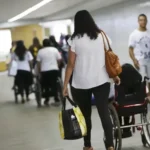 Corredor amplo de um espaço público, com várias pessoas circulando. Em destaque, uma mulher de cabelos médios e ondulados, vestindo blusa branca e calça preta, empurra uma pessoa em cadeira de rodas.