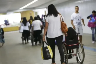 Corredor amplo de um espaço público, com várias pessoas circulando. Em destaque, uma mulher de cabelos médios e ondulados, vestindo blusa branca e calça preta, empurra uma pessoa em cadeira de rodas.