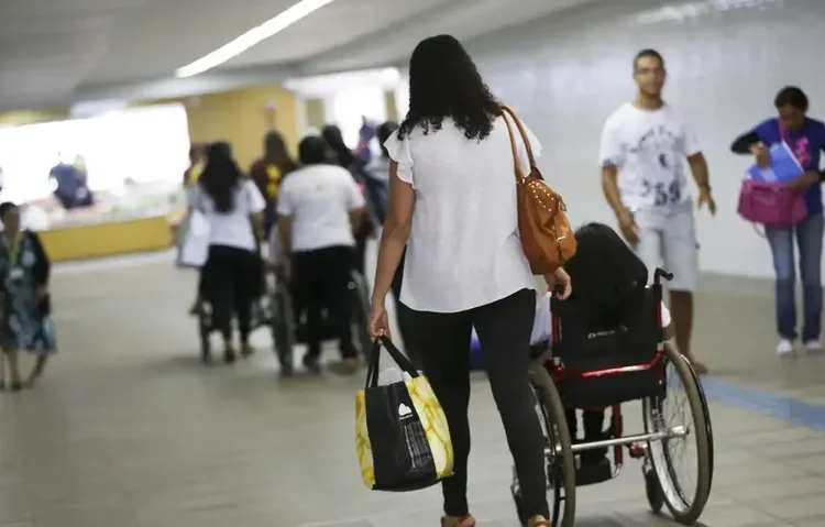 Corredor amplo de um espaço público, com várias pessoas circulando. Em destaque, uma mulher de cabelos médios e ondulados, vestindo blusa branca e calça preta, empurra uma pessoa em cadeira de rodas.