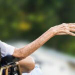 Idosa em cadeira de rodas com o braço estendido e a mão erguida. A pele frágil apresenta sinais de envelhecimento. Ao fundo, o cenário natural está desfocado, indicando que ela está ao ar livre.
