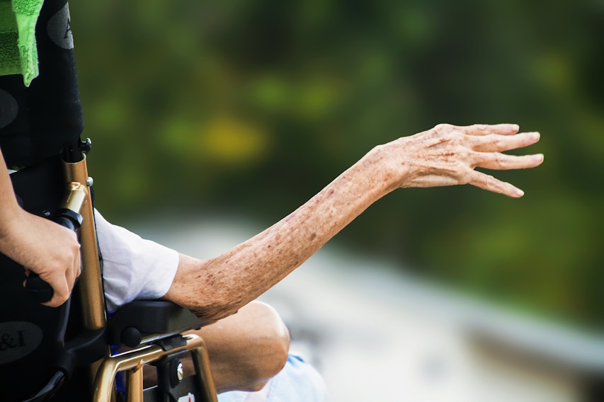 Idosa em cadeira de rodas com o braço estendido e a mão erguida. A pele frágil apresenta sinais de envelhecimento. Ao fundo, o cenário natural está desfocado, indicando que ela está ao ar livre.