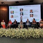 Pessoas sentadas em poltronas no palco durante a cerimônia de abertura do IX Fórum Ibero-Americano Inclui. Ao fundo, um telão exibe o nome do evento, fotos e nomes dos participantes. O palco está decorado com flores brancas e há uma bandeira ao lado direito. O ambiente é formal e voltado ao debate sobre inclusão social.