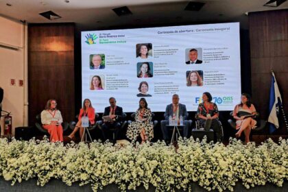Pessoas sentadas em poltronas no palco durante a cerimônia de abertura do IX Fórum Ibero-Americano Inclui. Ao fundo, um telão exibe o nome do evento, fotos e nomes dos participantes. O palco está decorado com flores brancas e há uma bandeira ao lado direito. O ambiente é formal e voltado ao debate sobre inclusão social.