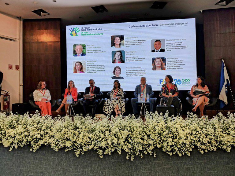 Pessoas sentadas em poltronas no palco durante a cerimônia de abertura do IX Fórum Ibero-Americano Inclui. Ao fundo, um telão exibe o nome do evento, fotos e nomes dos participantes. O palco está decorado com flores brancas e há uma bandeira ao lado direito. O ambiente é formal e voltado ao debate sobre inclusão social.