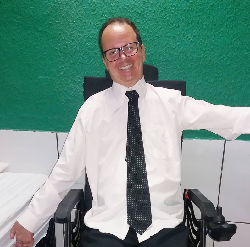 Sou o Angelo e estou sorrindo sentado em cadeira de rodas, vestindo camisa social branca, gravata preta e óculos, com fundo de parede verde.