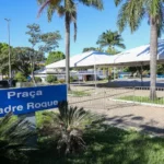 Vista da Praça Padre Roque durante o dia, com céu azul, árvores e palmeiras ao fundo. Há tendas brancas montadas e grades metálicas cercando parte da área. Em primeiro plano, uma placa azul com o nome da praça e arbustos ao redor.