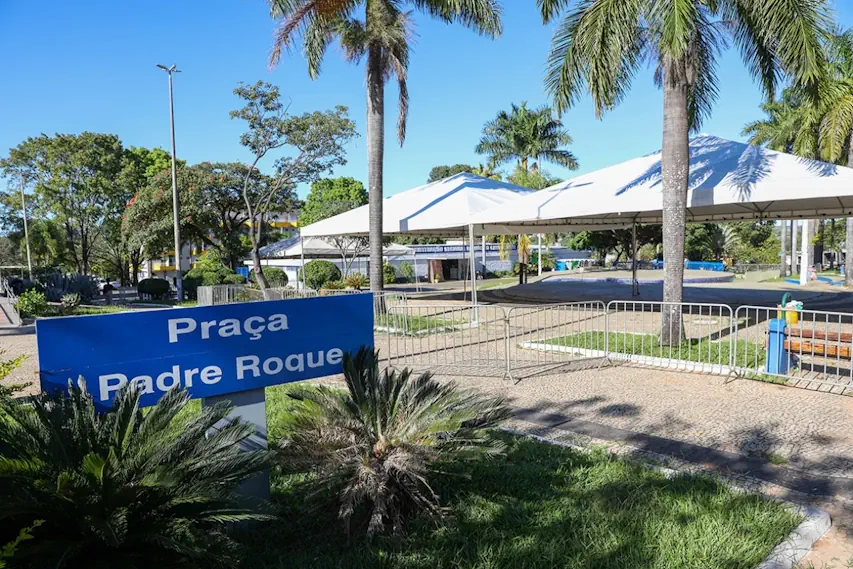 Vista da Praça Padre Roque durante o dia, com céu azul, árvores e palmeiras ao fundo. Há tendas brancas montadas e grades metálicas cercando parte da área. Em primeiro plano, uma placa azul com o nome da praça e arbustos ao redor.