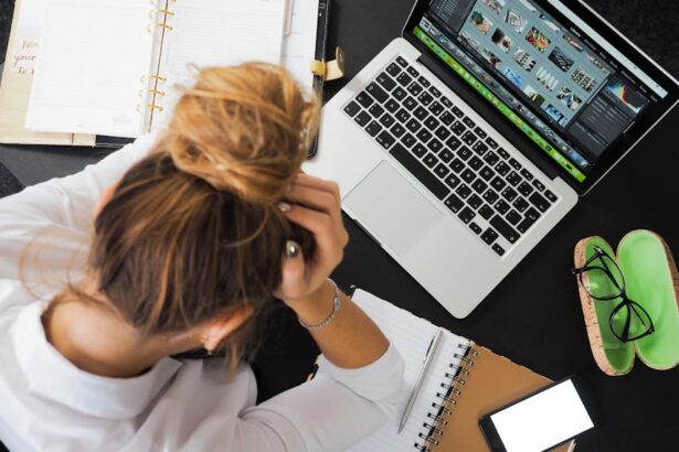 Pessoa com cabelo preso em um coque, vista de cima, com as mãos na cabeça em sinal de estresse ou cansaço. Ela está sentada à mesa, em frente a um notebook com imagens abertas na tela, ao lado de cadernos, celular, óculos e um estojo verde.