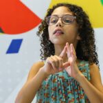 Criança de cabelos cacheados e óculos, usando vestido azul estampado com raposas, realiza sinal em Libras (Língua Brasileira de Sinais) com as mãos. Ao fundo, há um painel colorido com formas geométricas nas cores vermelho, azul, verde, amarelo, branco e preto.