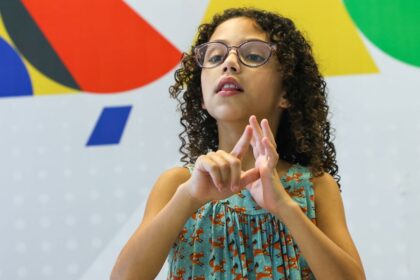 Criança de cabelos cacheados e óculos, usando vestido azul estampado com raposas, realiza sinal em Libras (Língua Brasileira de Sinais) com as mãos. Ao fundo, há um painel colorido com formas geométricas nas cores vermelho, azul, verde, amarelo, branco e preto.