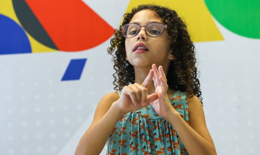 Criança de cabelos cacheados e óculos, usando vestido azul estampado com raposas, realiza sinal em Libras (Língua Brasileira de Sinais) com as mãos. Ao fundo, há um painel colorido com formas geométricas nas cores vermelho, azul, verde, amarelo, branco e preto.