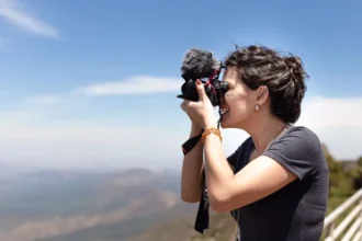 Mulher sorridente segurando uma câmera profissional com microfone acoplado, enquanto tira uma foto ao ar livre. Ela está em um ambiente montanhoso com céu azul ao fundo e usa camiseta cinza, brincos discretos e pulseiras coloridas. A paisagem atrás está desfocada, destacando a ação da fotografia.