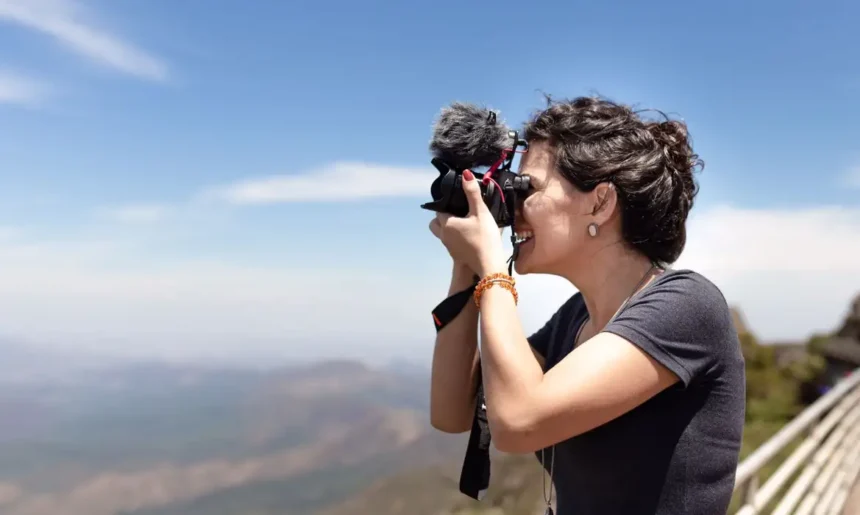 Mulher sorridente segurando uma câmera profissional com microfone acoplado, enquanto tira uma foto ao ar livre. Ela está em um ambiente montanhoso com céu azul ao fundo e usa camiseta cinza, brincos discretos e pulseiras coloridas. A paisagem atrás está desfocada, destacando a ação da fotografia.