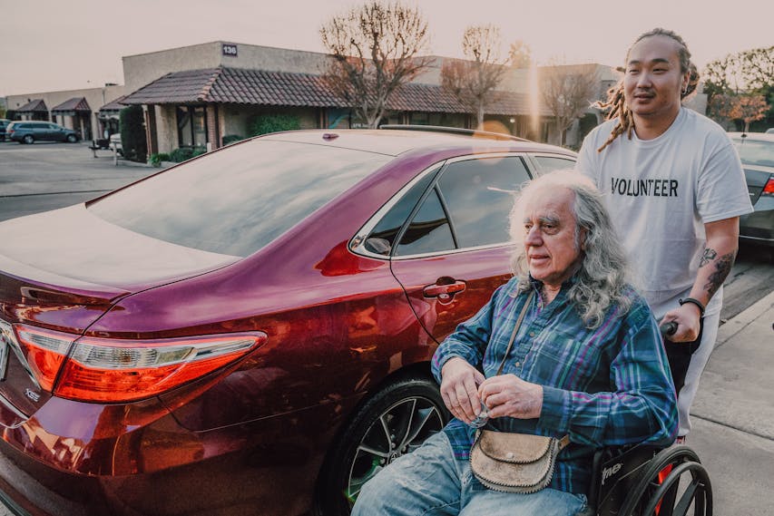 Um homem idoso com cabelos longos e grisalhos está sentado em uma cadeira de rodas ao lado de um carro vermelho, sendo empurrado por um jovem voluntário com cabelo preso e camiseta branca. Estão ao ar livre, em um estacionamento, com construções comerciais ao fundo e árvores secas ao pôr do sol. - Idosos-e-pessoas-com-deficiencia