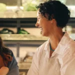 Duas mulheres sorridentes, uma com síndrome de Down, em um ambiente que parece ser um mercado ou loja de produtos frescos. A mulher com síndrome de Down está usando um avental marrom sobre uma camisa jeans azul, com os braços cruzados e os olhos fechados enquanto ri. A outra mulher, com cabelo encaracolado escuro e vestindo um avental branco, está olhando para ela e sorrindo. Há prateleiras com plantas verdes desfocadas ao fundo.