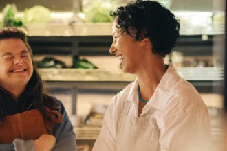Duas mulheres sorridentes, uma com síndrome de Down, em um ambiente que parece ser um mercado ou loja de produtos frescos. A mulher com síndrome de Down está usando um avental marrom sobre uma camisa jeans azul, com os braços cruzados e os olhos fechados enquanto ri. A outra mulher, com cabelo encaracolado escuro e vestindo um avental branco, está olhando para ela e sorrindo. Há prateleiras com plantas verdes desfocadas ao fundo.