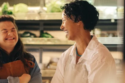 Duas mulheres sorridentes, uma com síndrome de Down, em um ambiente que parece ser um mercado ou loja de produtos frescos. A mulher com síndrome de Down está usando um avental marrom sobre uma camisa jeans azul, com os braços cruzados e os olhos fechados enquanto ri. A outra mulher, com cabelo encaracolado escuro e vestindo um avental branco, está olhando para ela e sorrindo. Há prateleiras com plantas verdes desfocadas ao fundo.
