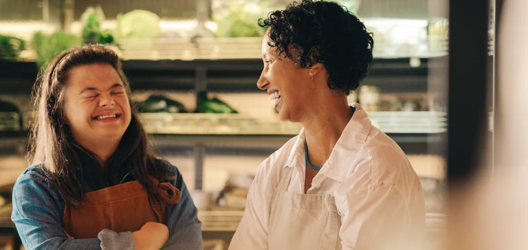 Duas mulheres sorridentes, uma com síndrome de Down, em um ambiente que parece ser um mercado ou loja de produtos frescos. A mulher com síndrome de Down está usando um avental marrom sobre uma camisa jeans azul, com os braços cruzados e os olhos fechados enquanto ri. A outra mulher, com cabelo encaracolado escuro e vestindo um avental branco, está olhando para ela e sorrindo. Há prateleiras com plantas verdes desfocadas ao fundo.