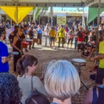 Parada do Orgulho da Pessoa com Deficiência, em Belo Horizonte Grupo diverso de pessoas se reúne em círculo sob uma tenda ao ar livre durante um evento cultural. No centro, músicos tocam instrumentos de percussão como surdos, tamborins e agogôs, enquanto o público observa ao redor. A atmosfera é vibrante e inclusiva. Ao fundo, há mais espectadores e barracas com faixas e placas coloridas. O evento ocorre em um espaço público pavimentado com pedras.