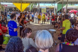 Parada do Orgulho da Pessoa com Deficiência, em Belo Horizonte Grupo diverso de pessoas se reúne em círculo sob uma tenda ao ar livre durante um evento cultural. No centro, músicos tocam instrumentos de percussão como surdos, tamborins e agogôs, enquanto o público observa ao redor. A atmosfera é vibrante e inclusiva. Ao fundo, há mais espectadores e barracas com faixas e placas coloridas. O evento ocorre em um espaço público pavimentado com pedras.