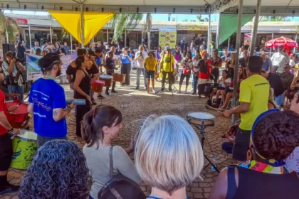 Parada do Orgulho da Pessoa com Deficiência, em Belo Horizonte Grupo diverso de pessoas se reúne em círculo sob uma tenda ao ar livre durante um evento cultural. No centro, músicos tocam instrumentos de percussão como surdos, tamborins e agogôs, enquanto o público observa ao redor. A atmosfera é vibrante e inclusiva. Ao fundo, há mais espectadores e barracas com faixas e placas coloridas. O evento ocorre em um espaço público pavimentado com pedras.
