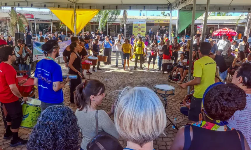 Parada do Orgulho da Pessoa com Deficiência, em Belo Horizonte Grupo diverso de pessoas se reúne em círculo sob uma tenda ao ar livre durante um evento cultural. No centro, músicos tocam instrumentos de percussão como surdos, tamborins e agogôs, enquanto o público observa ao redor. A atmosfera é vibrante e inclusiva. Ao fundo, há mais espectadores e barracas com faixas e placas coloridas. O evento ocorre em um espaço público pavimentado com pedras.