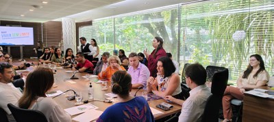 Em uma ampla sala de reuniões com janelas de vidro e vista para um jardim, dezenas de pessoas estão reunidas ao redor de uma grande mesa de madeira. Uma mulher de pé fala ao grupo, enquanto os demais escutam atentamente. À esquerda da imagem, uma tela mostra o título “Novo Viver Sem Limite”, relacionado ao Plano Nacional dos Direitos da Pessoa com Deficiência. O ambiente é iluminado e transmite formalidade e cooperação.