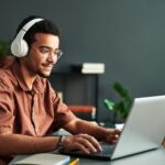 Jovem com deficiência auditiva, usando fones de ouvido grandes e óculos, trabalha concentrado em um notebook. Ele está sentado em uma mesa com livros e plantas ao fundo, em um ambiente interno e acolhedor.