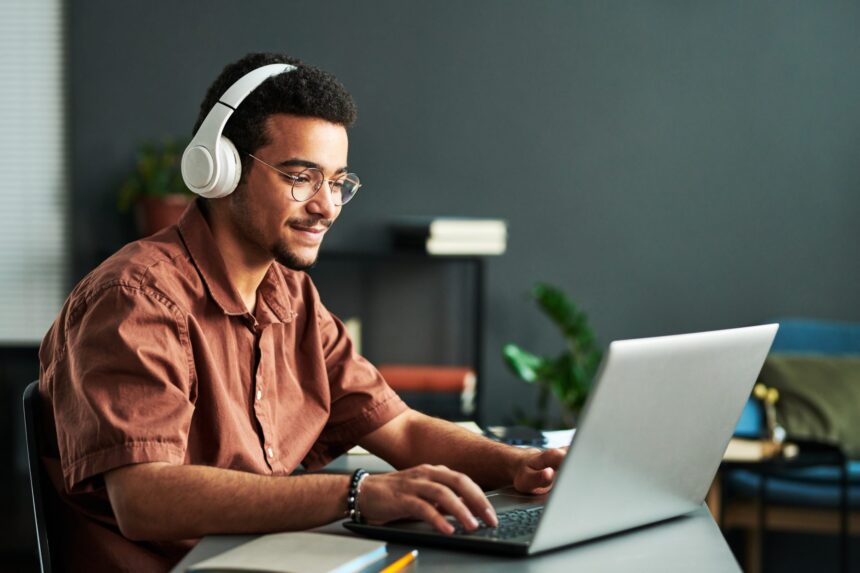 Jovem com deficiência auditiva, usando fones de ouvido grandes e óculos, trabalha concentrado em um notebook. Ele está sentado em uma mesa com livros e plantas ao fundo, em um ambiente interno e acolhedor.