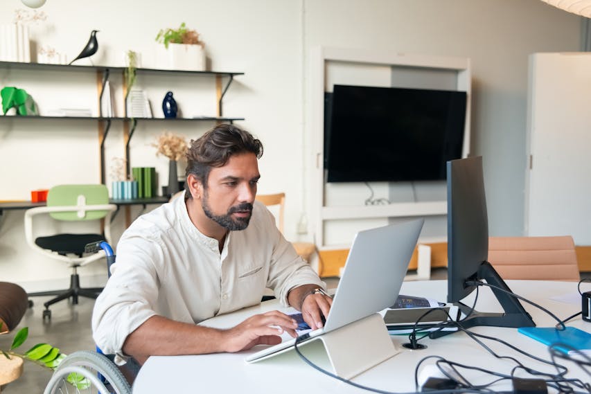 Homem cadeirante, com barba e cabelo castanho escuro, trabalha concentrado em um notebook em uma sala moderna. Ele usa uma camisa clara e está cercado por monitores e cabos sobre a mesa. Ao fundo, há prateleiras decoradas e uma televisão. A imagem representa inclusão e protagonismo/empreendedorismo da pessoa com deficiência no ambiente profissional.