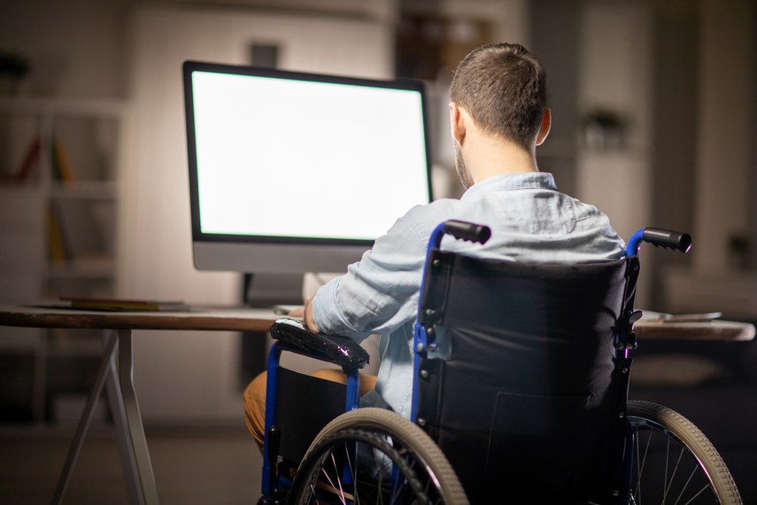 Homem cadeirante, de costas, trabalha em frente a um computador de tela grande e branca. Ele está em um ambiente interno, sentado à mesa, usando uma camisa clara. A cena representa autonomia e inclusão digital, podendo estar associada ao empreendedorismo de pessoas com deficiência.
