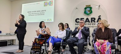 Em um auditório oficial do Governo do Ceará, seis pessoas estão sentadas em cadeiras sobre um palco. À esquerda, uma intérprete de Libras traduz o evento. No centro do painel, um telão exibe o título: “Novo Viver sem Limite – Plano Nacional dos Direitos da Pessoa com Deficiência”. À direita da imagem, o brasão do Ceará está fixado na parede com os dizeres: "Governo do Estado do Ceará – Secretaria dos Direitos Humanos".