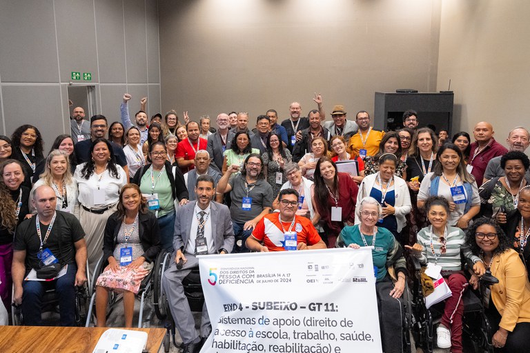 Grupo diverso de pessoas, incluindo pessoas com deficiência e usuários de cadeira de rodas, posa sorridente para a foto durante o 5º Fórum Nacional dos Direitos da Pessoa com Deficiência, realizado em Brasília, de 14 a 17 de julho de 2024. Ao centro, uma faixa destaca o tema do grupo de trabalho: “Sistemas de apoio – direito de acesso à escola, trabalho, saúde, habilitação, reabilitação”.