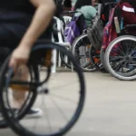 Grupo de pessoas com deficiência usando cadeiras de rodas reunidas em um espaço aberto. Algumas cadeiras possuem mochilas penduradas, sugerindo um ambiente de encontro ou evento. No primeiro plano, uma pessoa em cadeira de rodas manual aparece desfocada.
