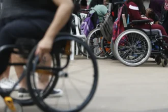 Grupo de pessoas com deficiência usando cadeiras de rodas reunidas em um espaço aberto. Algumas cadeiras possuem mochilas penduradas, sugerindo um ambiente de encontro ou evento. No primeiro plano, uma pessoa em cadeira de rodas manual aparece desfocada.
