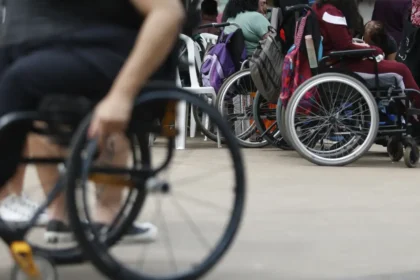 Grupo de pessoas com deficiência usando cadeiras de rodas reunidas em um espaço aberto. Algumas cadeiras possuem mochilas penduradas, sugerindo um ambiente de encontro ou evento. No primeiro plano, uma pessoa em cadeira de rodas manual aparece desfocada.